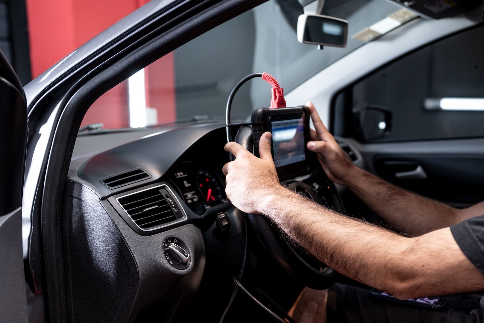An automotive technician using a diagnostic tool mounted on the steering wheel inside the modern interior of a car, checking technical data and performing system tests on the vehicle.