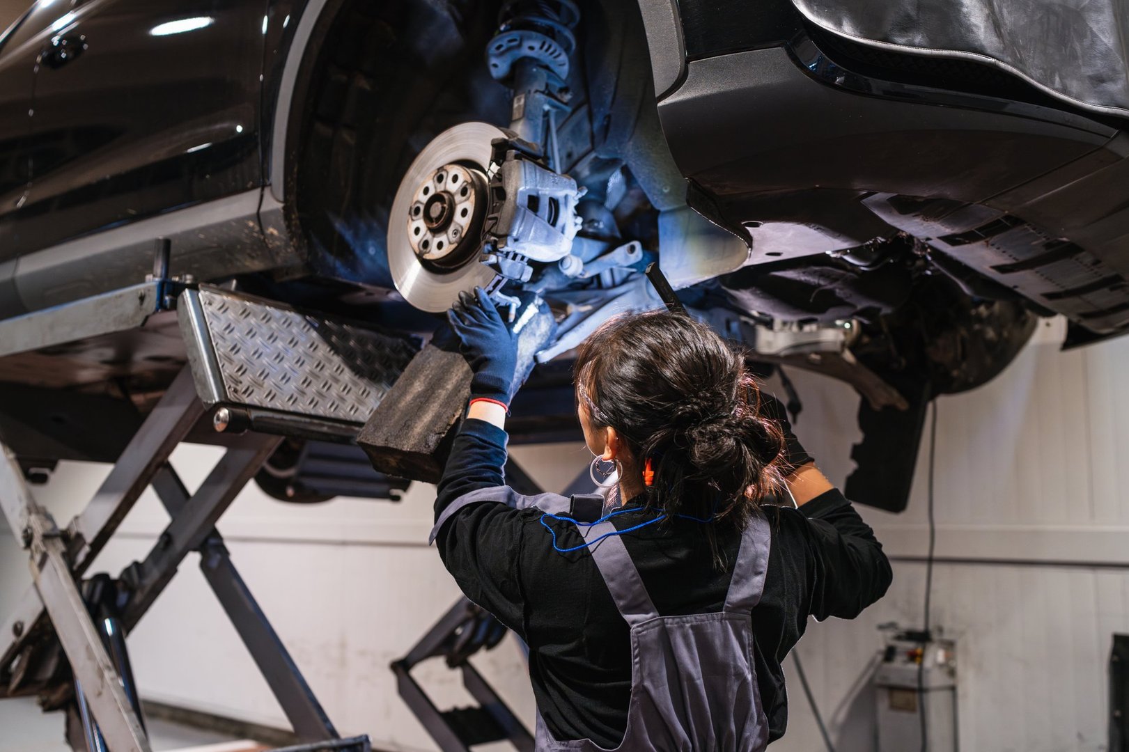 Female mechanic working under a car lifted on a hydraulic lift, inspecting brakes in a professional car repair shop